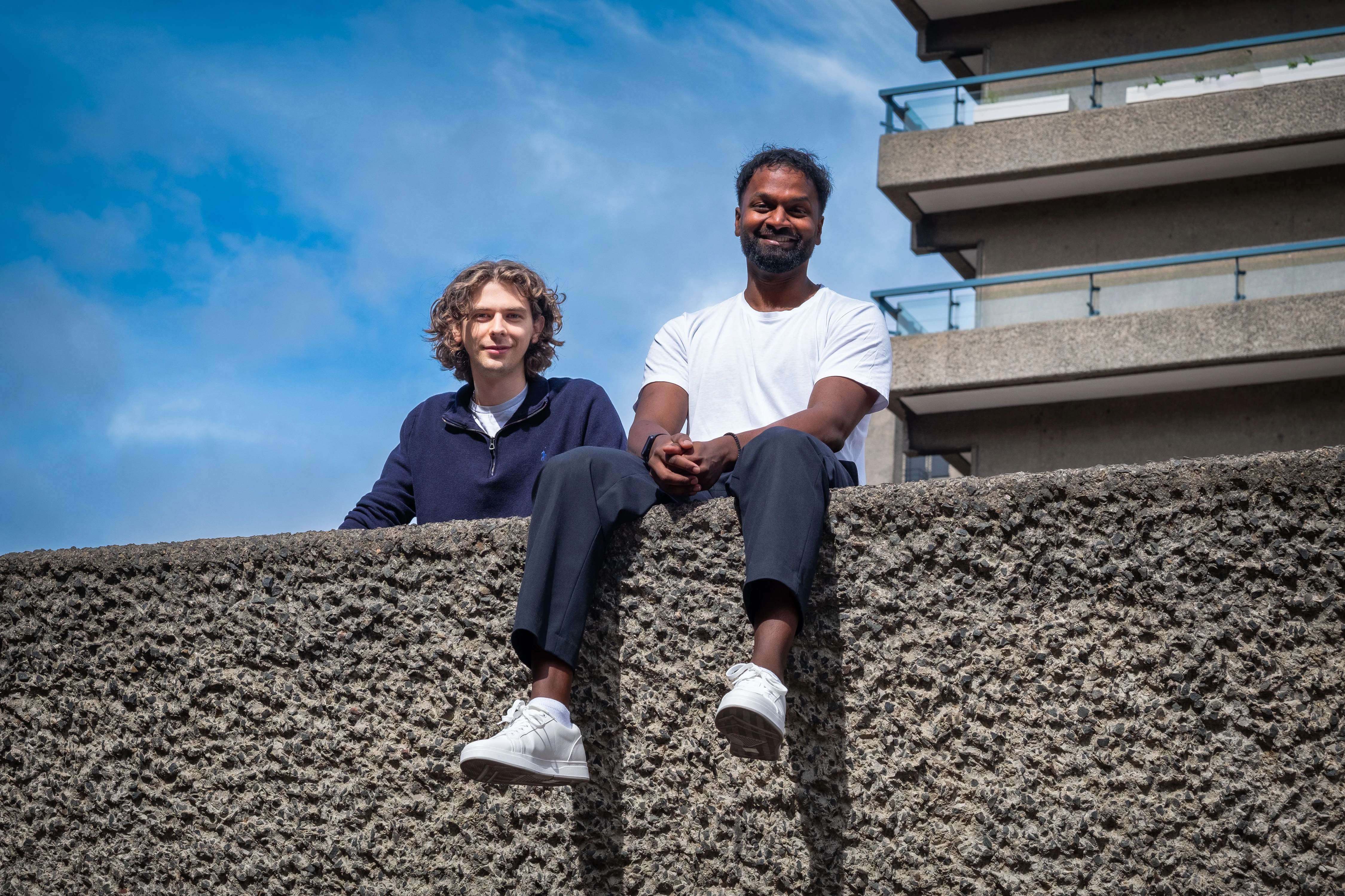 Zenithon AI Founders - Both founders seated on a Barbican concrete wall against blue sky, low angle